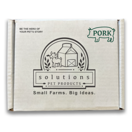A square cardboard box for Solutions Pet Products, featuring a cream-colored background with black and green printed graphics and text, including a pig logo and a farm illustration.